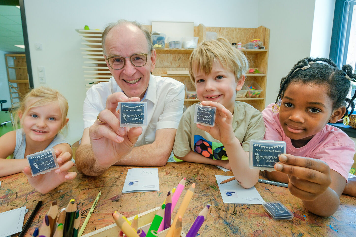 Eine männliche Person sitzt neben Kindern an einem Tisch mit Malsachen. Alle halten ein kleines, blaues Kästchen in Händen.