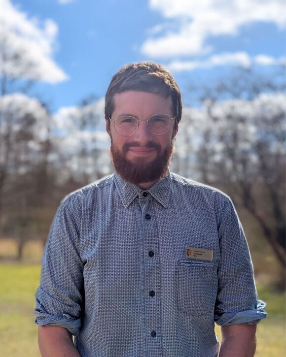 Eine männlich gelesene Person mit Bart und Brille steht in einem karierten Hemd im Grünen.